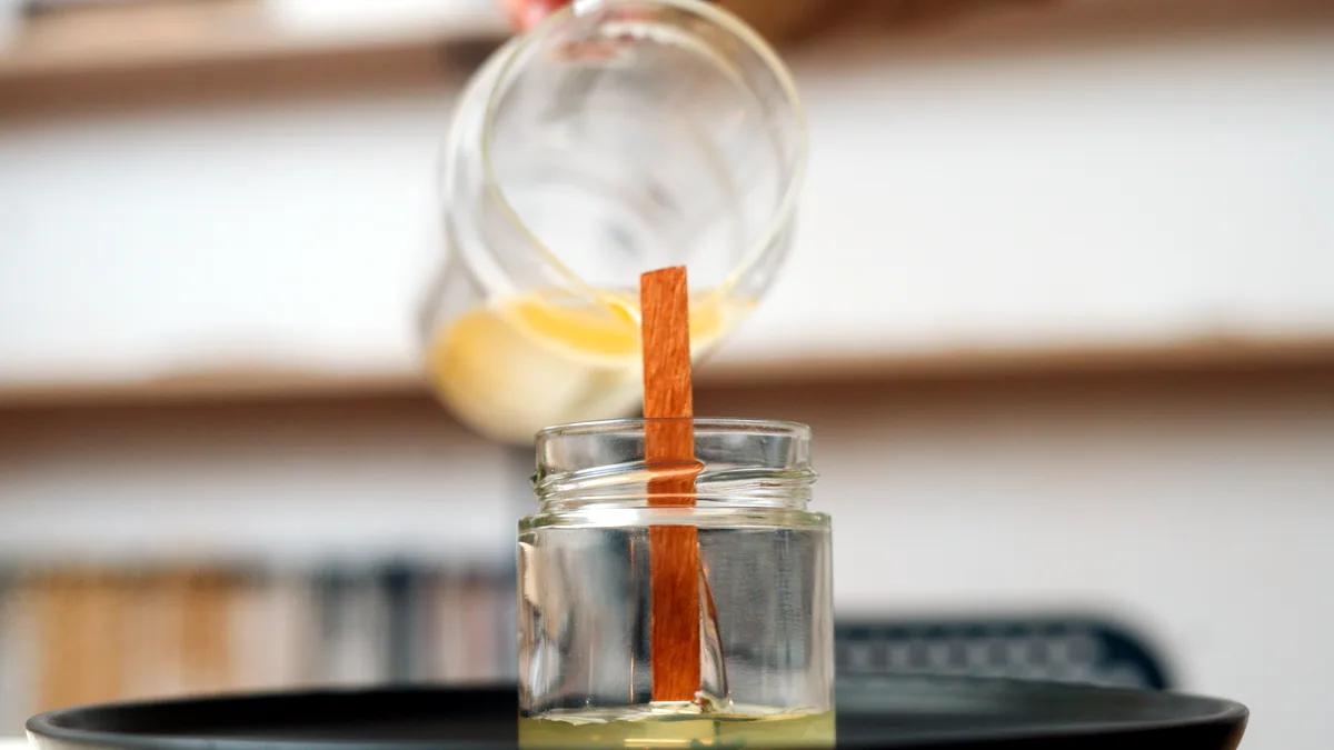 Amber wax being poured into a clear glass jar on a workbench.