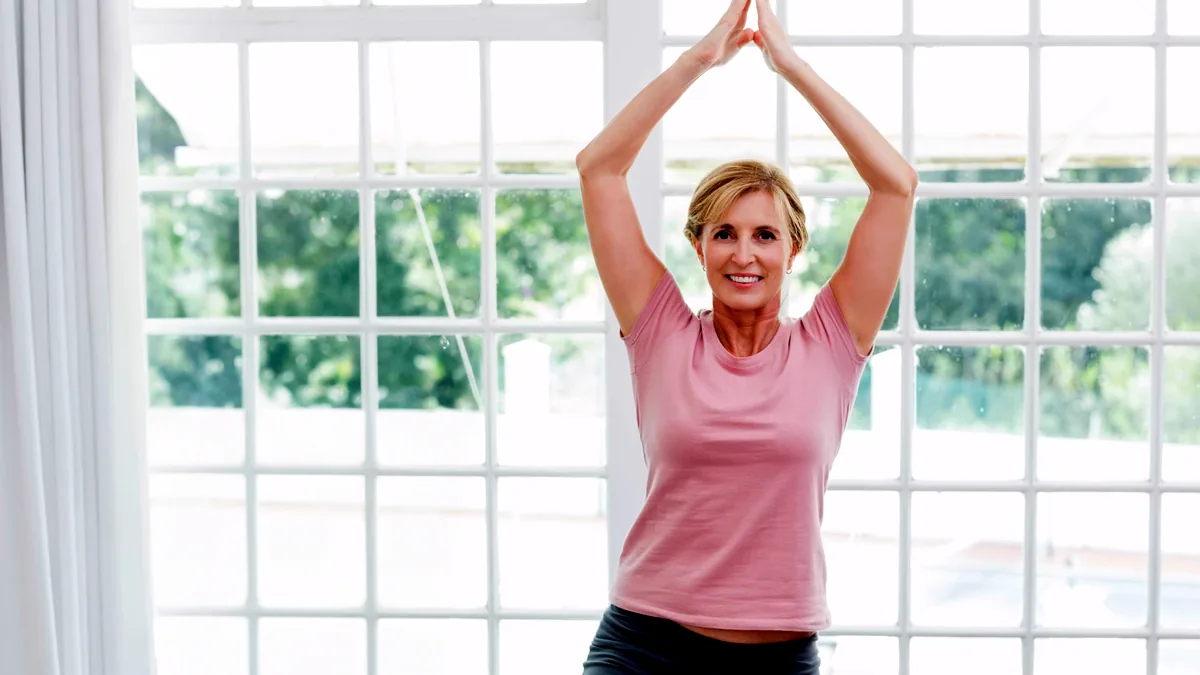 An older person standing by a bright window, arms raised in a stretch.